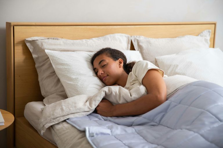 Person sleeping peacefully in a bright, calm bedroom, resting under a light blanket with soft pillows and a wooden headboard.