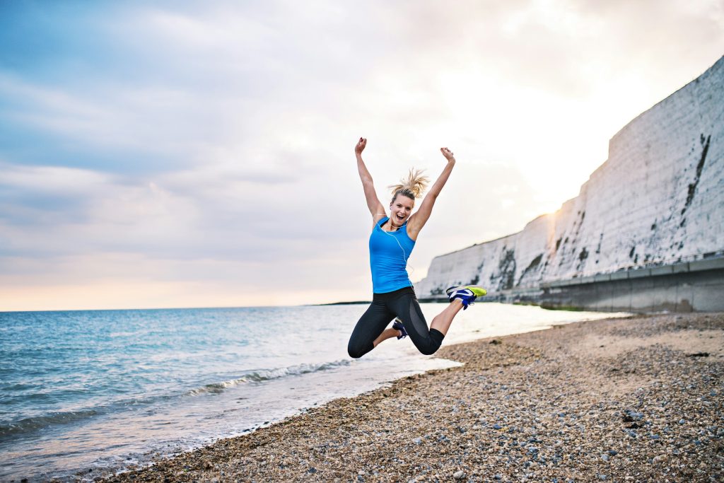 Woman in athletic clothing jumping joyfully on a beach near the water, suggesting energy, vitality, and improved exercise capacity.