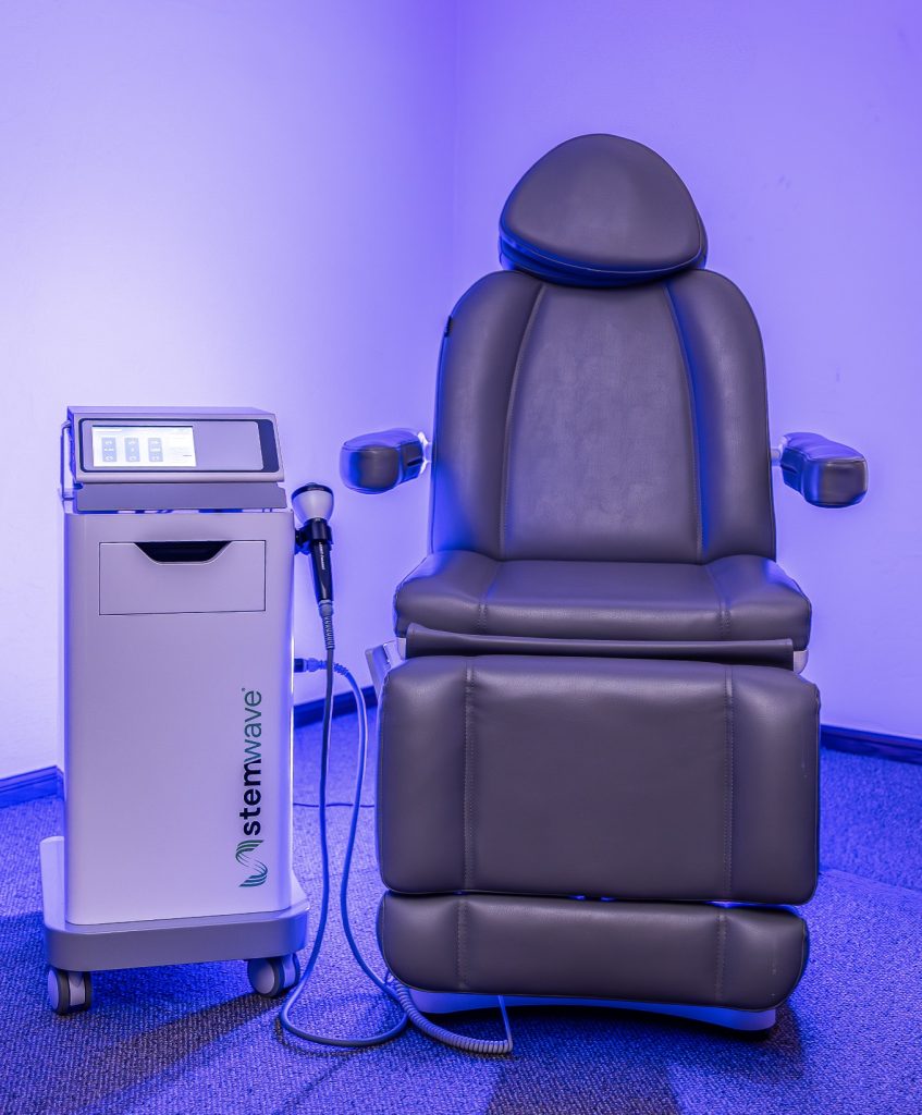StemWave shockwave therapy system and treatment chair at OK Theta & Wellness, shown in a blue-lit therapy room with the handheld acoustic wave applicator beside the chair.
