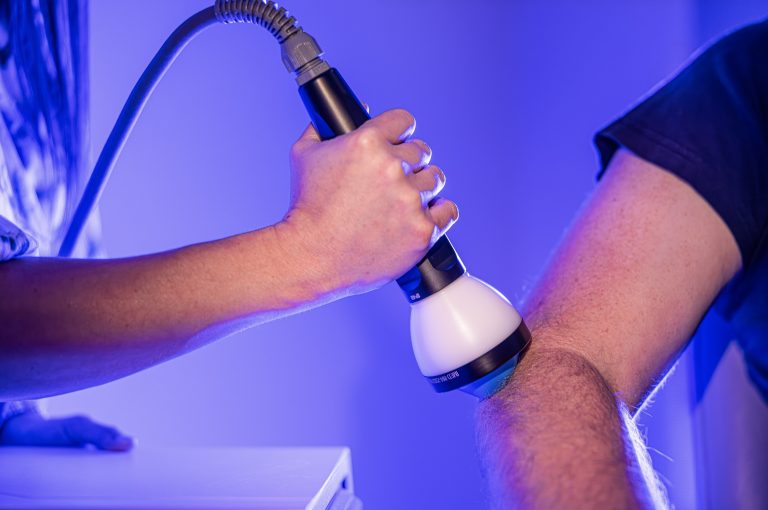 StemWave shockwave therapy being applied to a patient’s knee at OK Theta & Wellness, with a handheld acoustic wave device used over the joint area in blue clinical lighting.