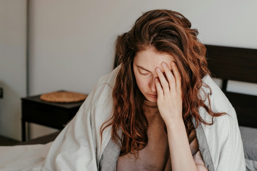 Woman sitting on a bed with her hand over her face, appearing exhausted or overwhelmed, illustrating fatigue, brain fog, low energy, or post-viral depletion.