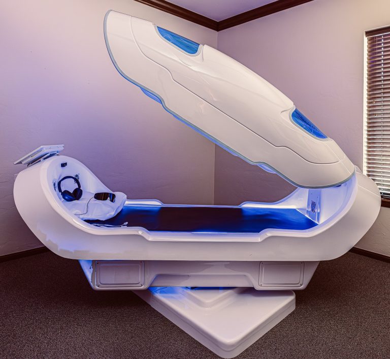 Open white Theta Chamber at OK Theta & Wellness with blue accent lighting, headphones, light glasses, and a padded interior prepared for a relaxation session.