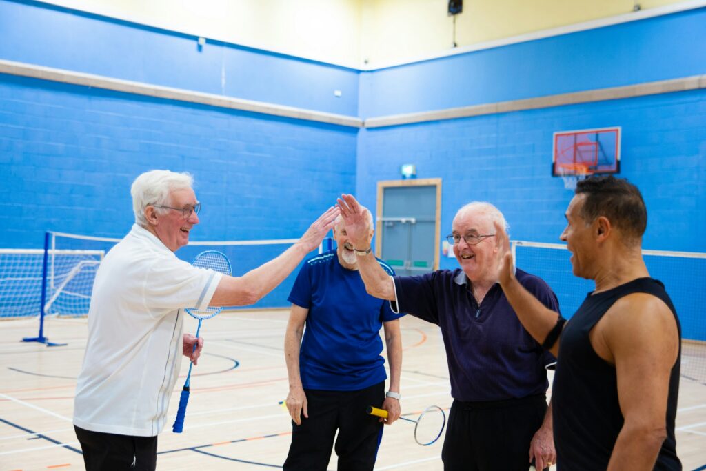 Four adults in a gym smile and high-five after playing racket sports, representing active aging, mobility, confidence, and staying engaged in life.