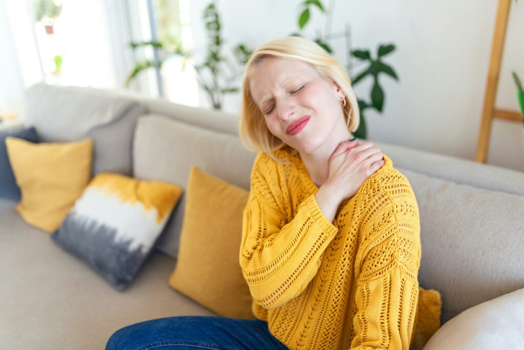 Woman sitting on a couch holding her neck and shoulder with a pained expression, representing chronic neck, shoulder, or soft tissue pain.