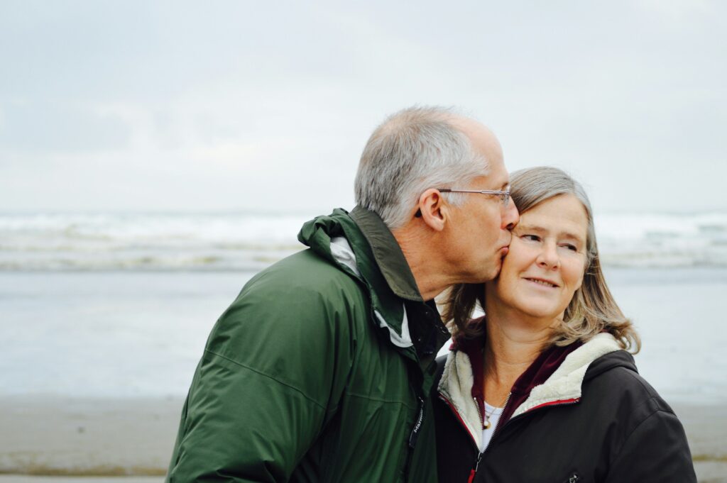 Older couple standing together on a beach, with one person kissing the other on the temple, representing active aging, companionship, mobility, and meaningful quality of life.