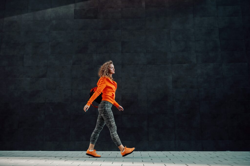 Woman in an orange sweatshirt walking confidently in front of a dark wall, representing improved movement, mobility, and function.