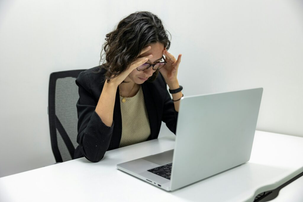 Person sitting at a desk with a laptop, holding their temples and looking fatigued or stressed.